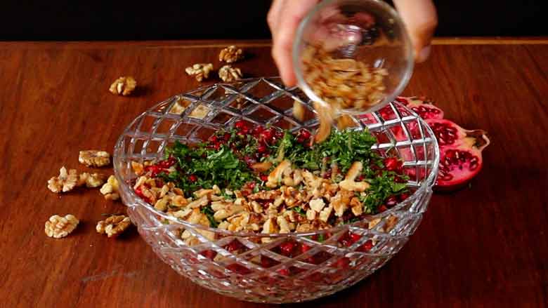 A hand is seen tipping toasted California walnuts from a glass bowl over the salad in large cut-glass bowl. California walnuts kernels and an open pomegranate lie on the wooden counter in the background - Konkani Cucumber and Pomegranate Salad Recipe